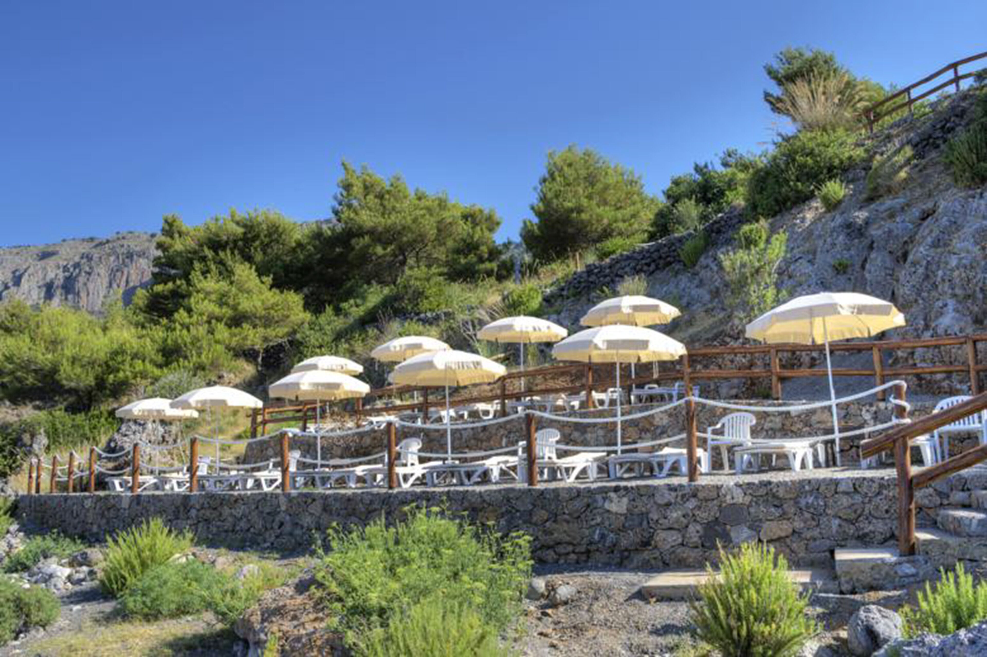 A terrace with outdoor seating and umbrellas, surrounded by greenery and a stone wall.