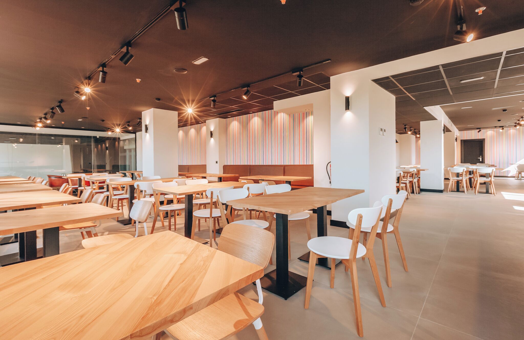 A spacious dining area with wooden tables and white chairs, featuring a modern design with striped wall panels and recessed lighting.