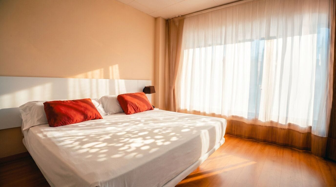 A hotel room with a large bed, a headboard, and a window with sheer curtains.