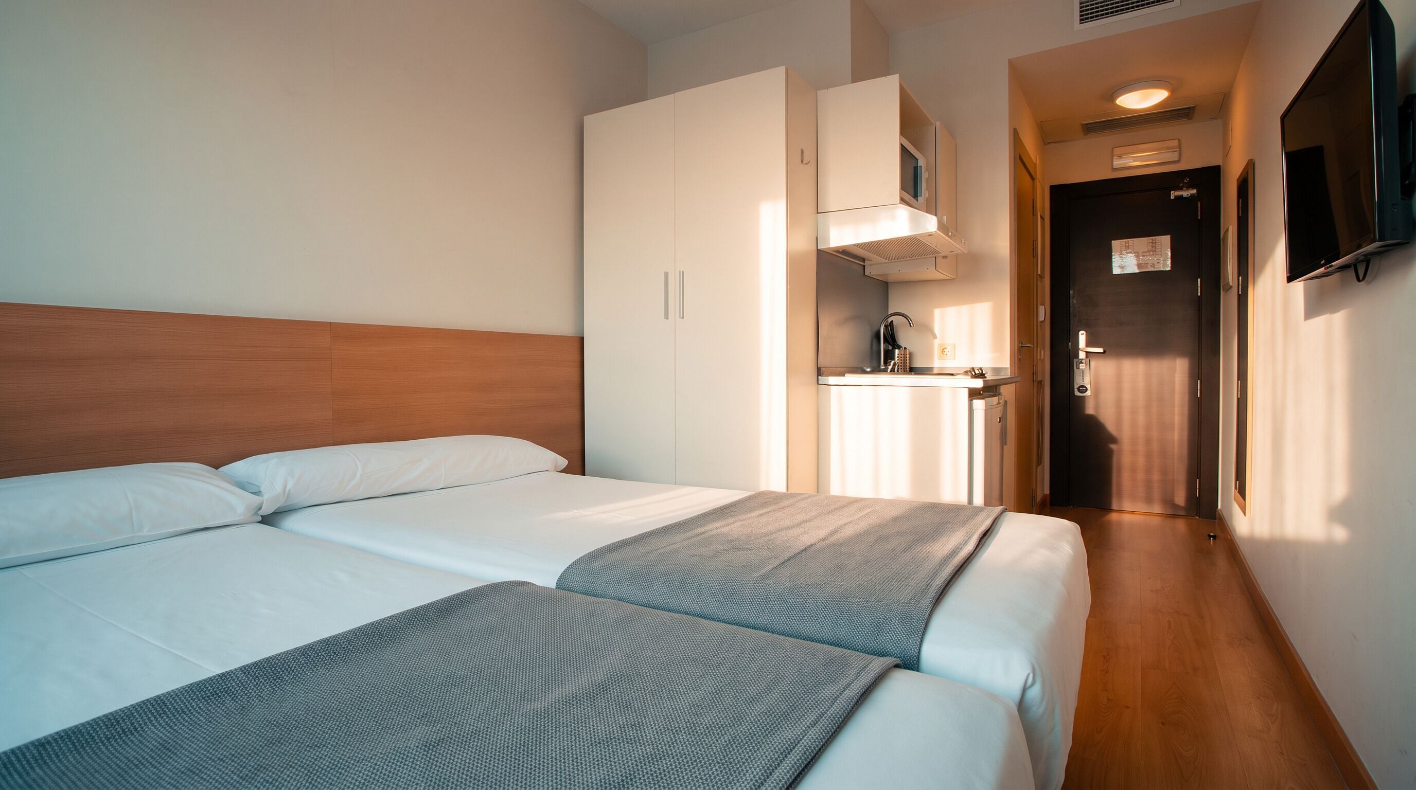 Una habitación de hotel con cabecera de madera, dos camas, una kitchenette y un televisor montado en la pared.