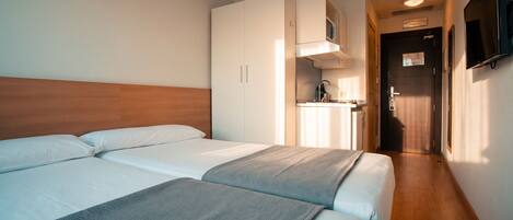 Una habitación de hotel con cabecera de madera, dos camas, una kitchenette y un televisor montado en la pared.