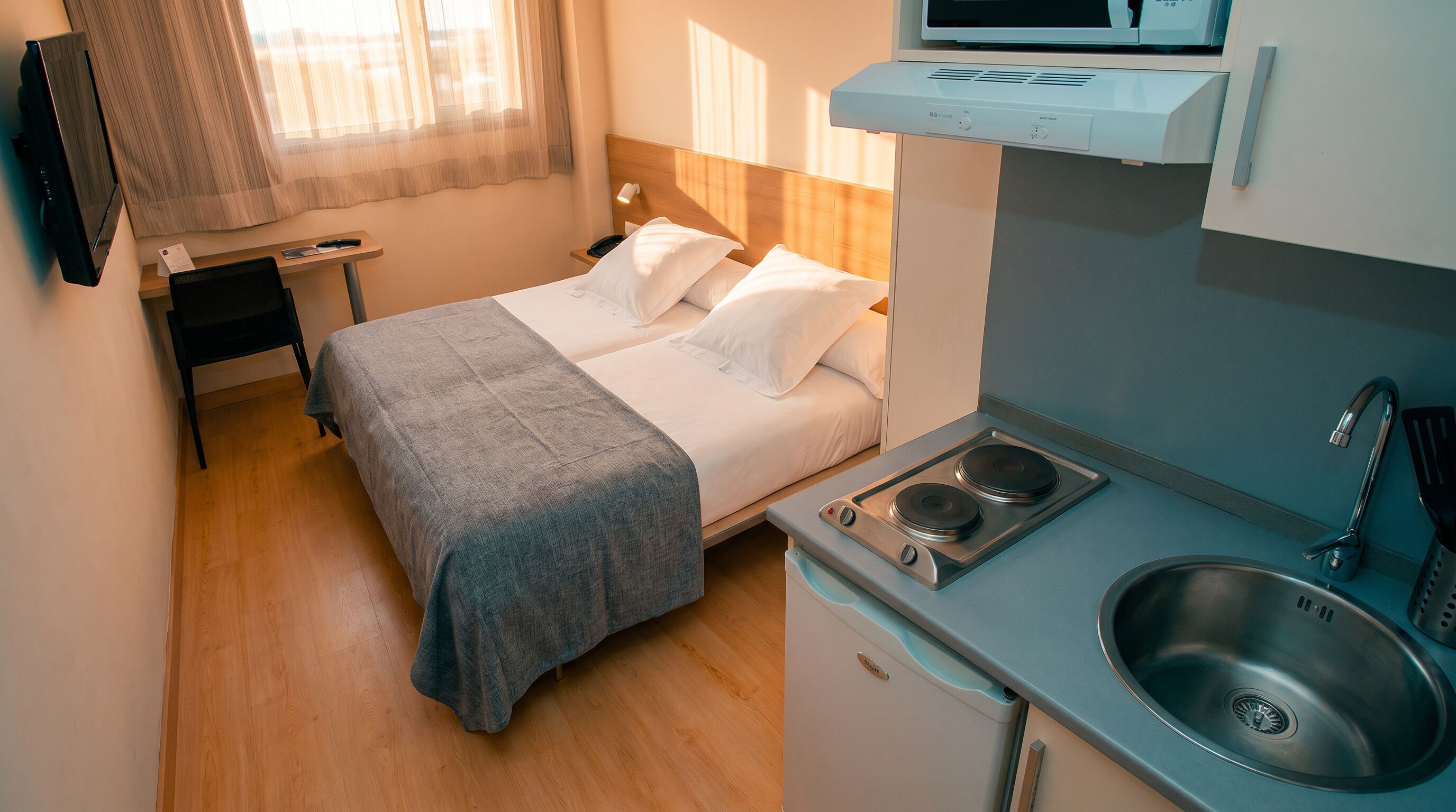 Et lille hotelværelse med seng, natbord, stol og et tekøkken med vask og komfur.
