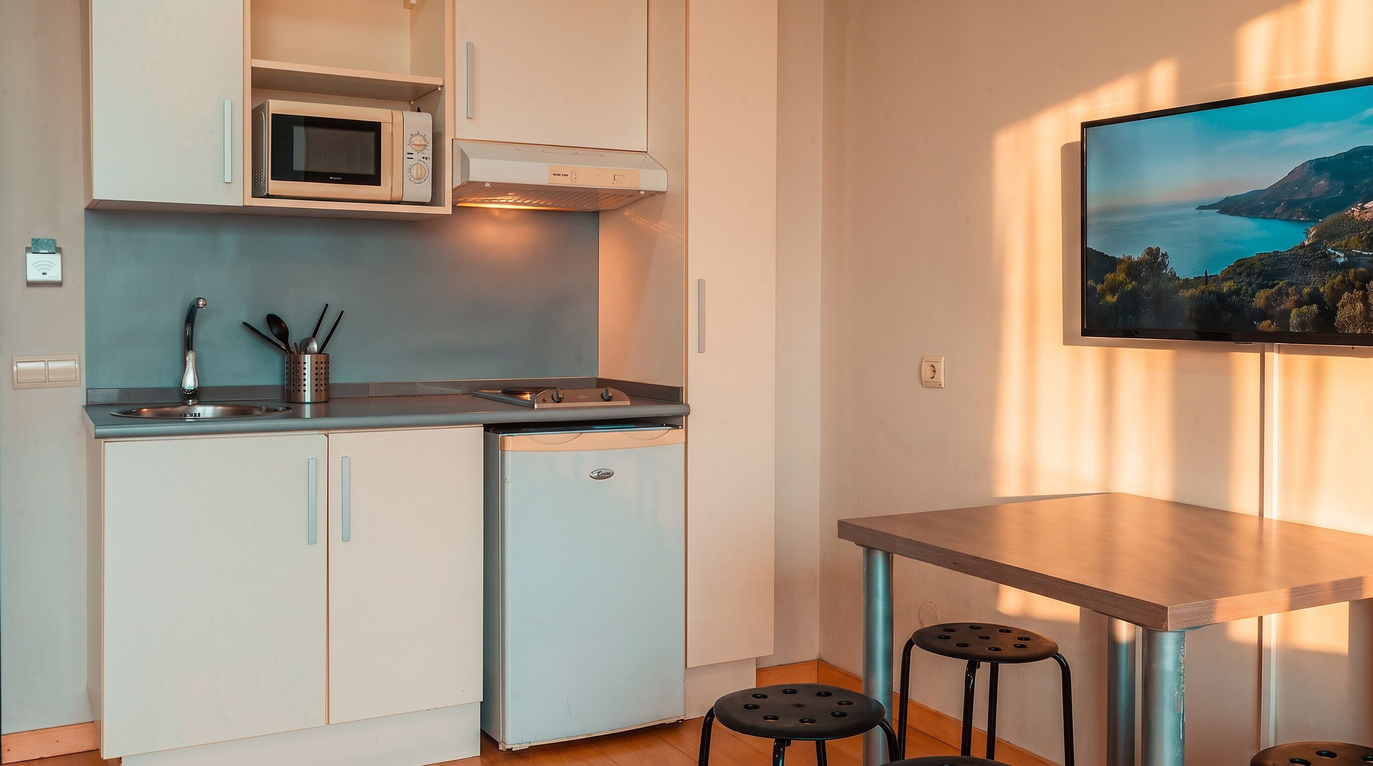 A compact kitchen with a microwave, stove, and sink, a small dining table with stools, and a TV mounted on the wall.