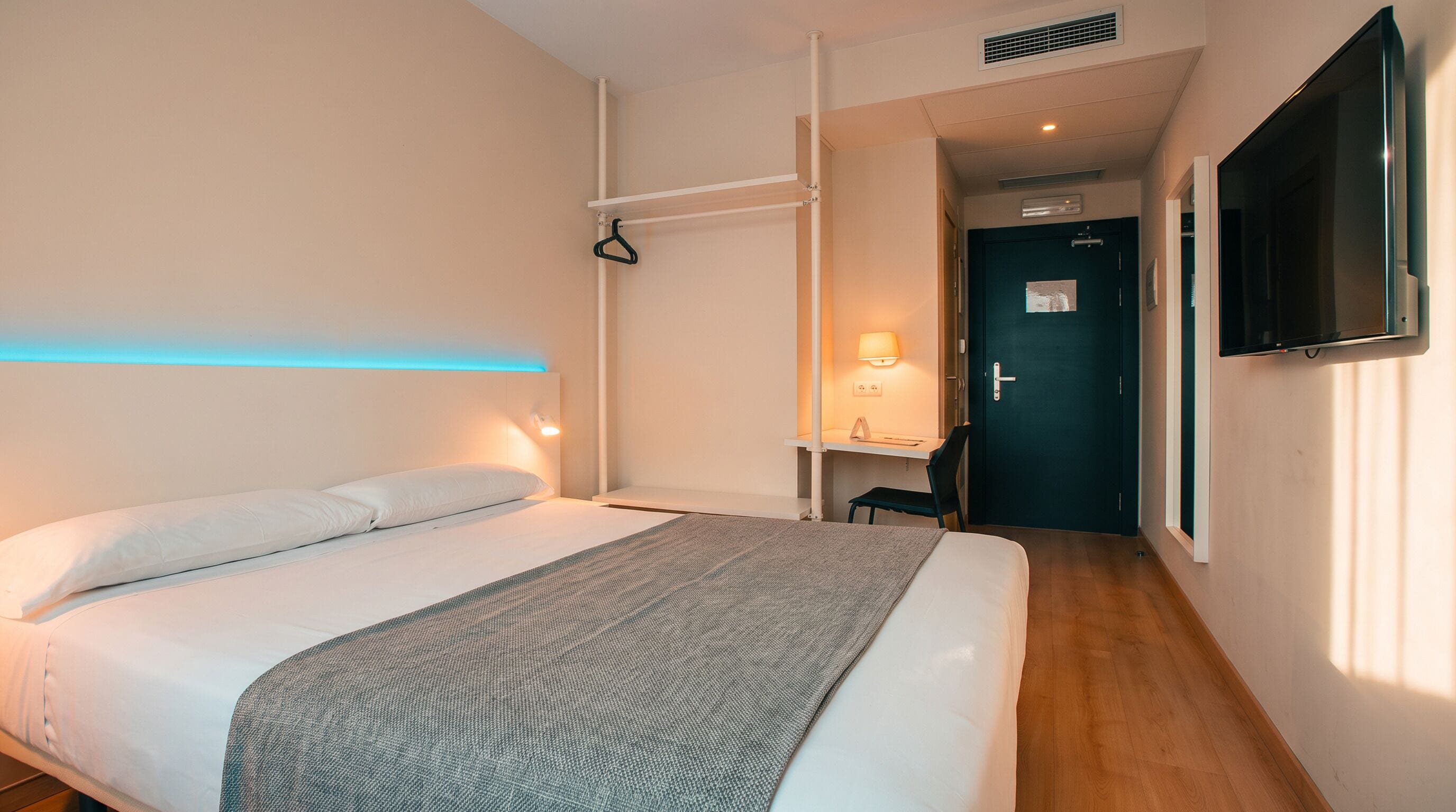 A hotel room with a bed, bedside table, lamp, and a TV mounted on the wall.