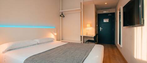 Una habitación de hotel con cama, mesita de noche, lámpara y televisor montado en la pared.