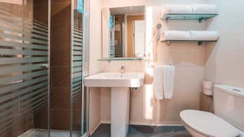 Un baño con mampara de vidrio, un lavamanos blanco, un inodoro y repisas de vidrio con toallas.
