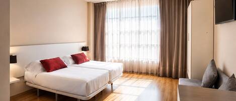 A hotel room with two beds, a wooden floor, a large window with curtains, a TV, and a desk.