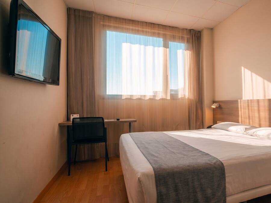 Habitación de hotel con cama, escritorio, silla, televisión y ventana con cortinas.