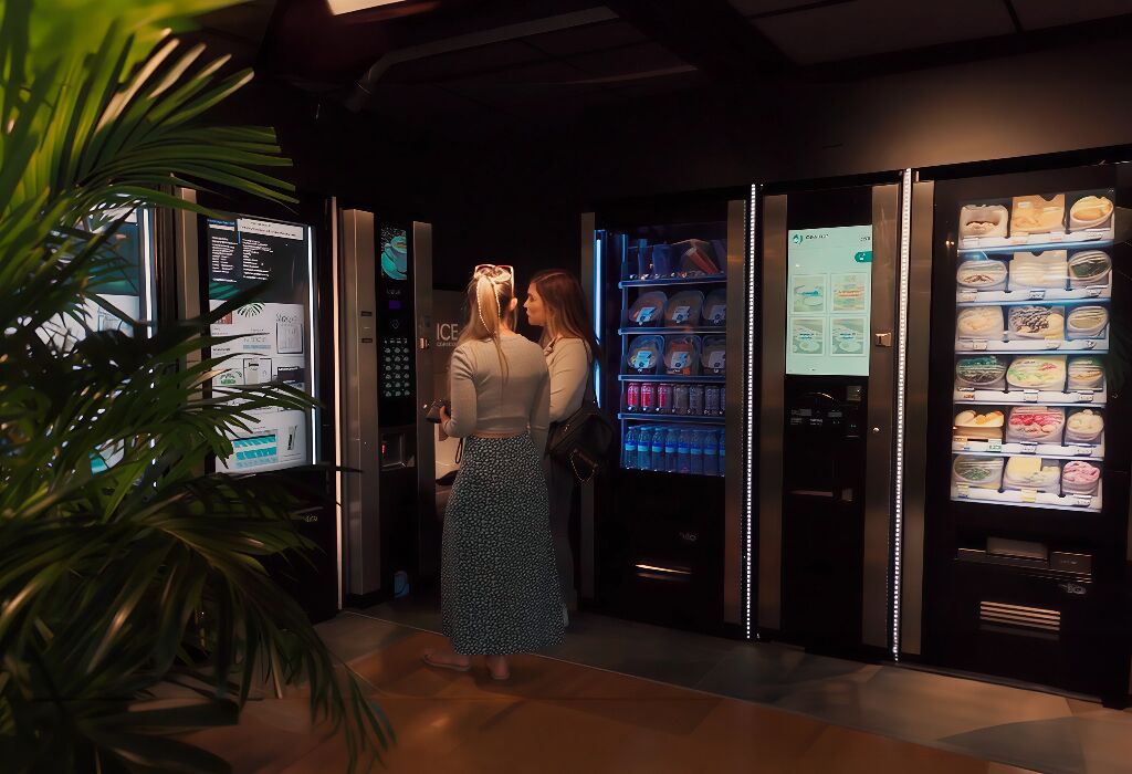 Un vestíbulo de hotel moderno con un quiosco de información digital, una máquina expendedora con diferentes productos alimenticios y una planta en maceta.