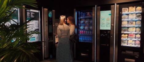 A modern hotel lobby with a digital information kiosk, a vending machine with various food items, and a potted plant.