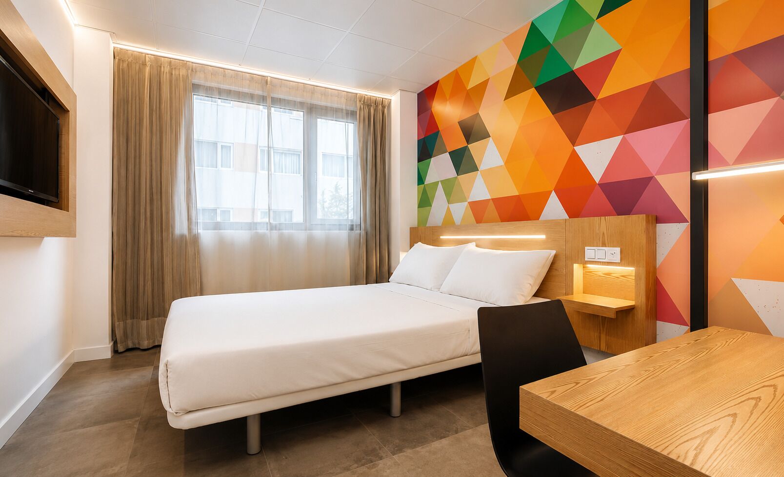 Habitación de hotel con cama, escritorio, silla, televisor montado en la pared y un llamativo diseño geométrico en la pared.