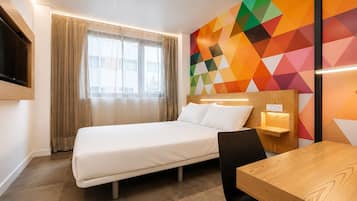 Habitación de hotel con cama, escritorio, silla, televisor montado en la pared y un llamativo diseño geométrico en la pared.