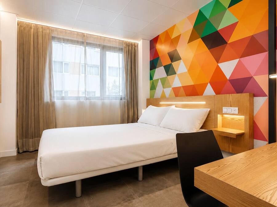Habitación de hotel con cama, escritorio, silla, televisor montado en la pared y un llamativo diseño geométrico en la pared.