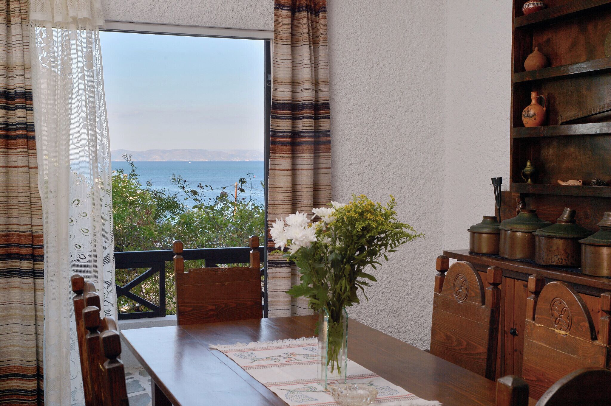 Un comedor con vista al mar, una mesa de madera, sillas, un florero con flores y una estantería con cerámica.