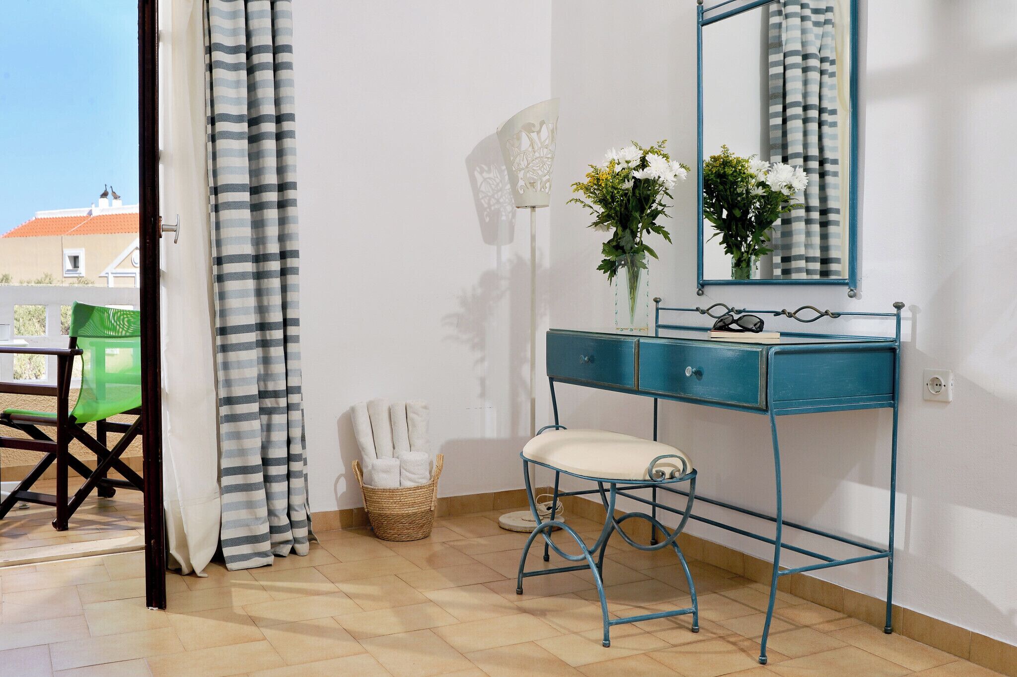 Una habitación con un tocador azul, un espejo, una cesta de mimbre con toallas, una silla verde y vistas a edificios.