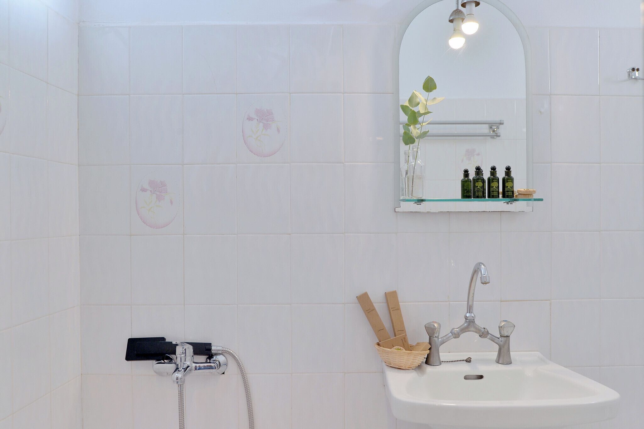 A bathroom with white tiles, a shower, a sink with a mirror, and a shelf with toiletries.