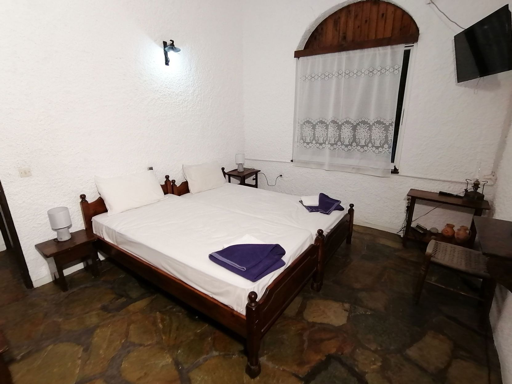 A bedroom with a stone floor, a wooden bed, a nightstand, a bedside table, a lamp, a window with a curtain, and a television on the wall.