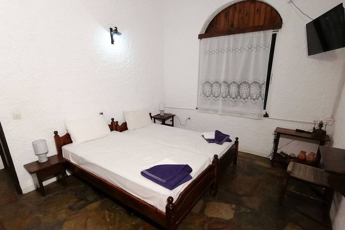 A bedroom with a stone floor, a wooden bed, a nightstand, a bedside table, a lamp, a window with a curtain, and a television on the wall.