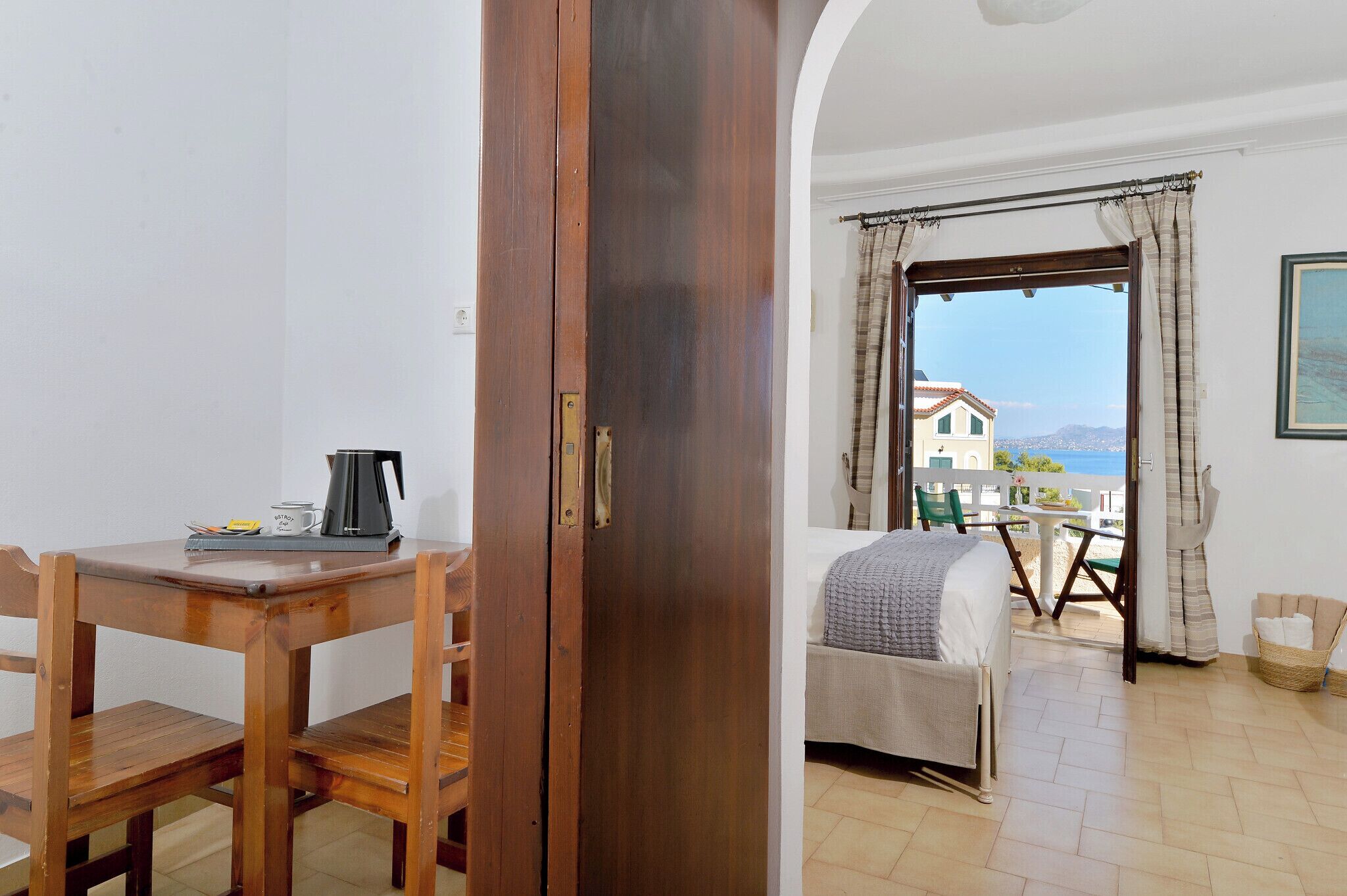 A room with a wooden table, a bed, and a view of the sea through a window with curtains.