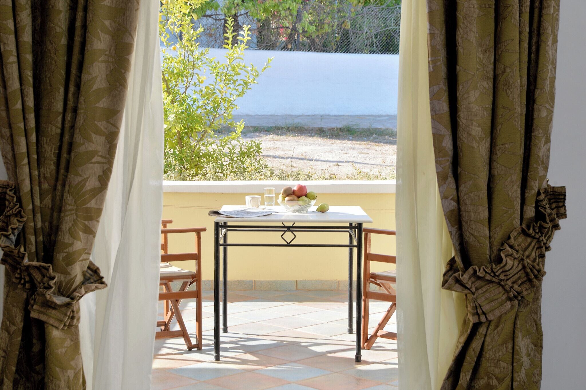 A table with a bowl of fruit and glasses, a window with a view of the sea, and curtains with a leaf pattern.