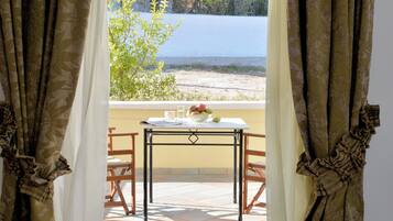 A table with a bowl of fruit and glasses, a window with a view of the sea, and curtains with a leaf pattern.