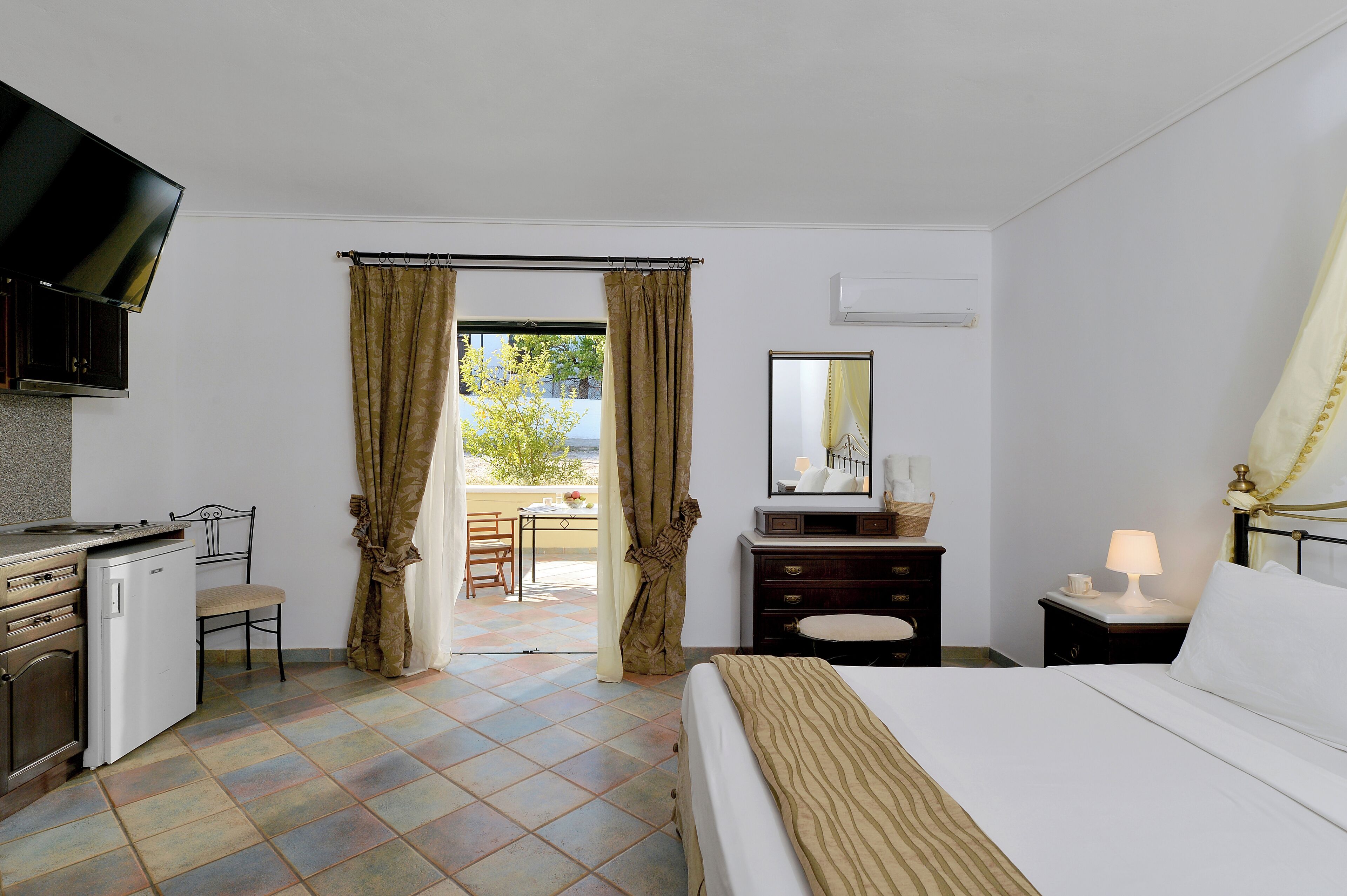 Una camera d'albergo con un letto, una scrivania, una televisione, un piccolo frigorifero, una sedia e una finestra con tende.