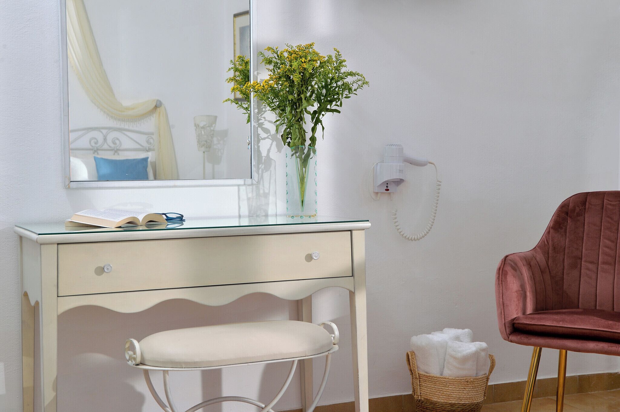 A room with a white desk, a chair, a vase with flowers, a mirror, a telephone, and a red chair.