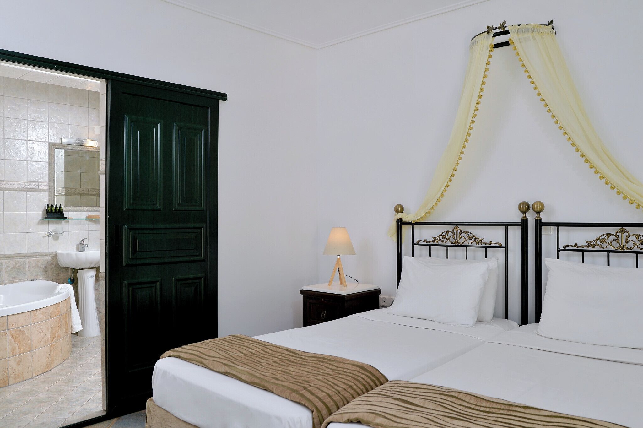 Habitación de hotel con cama, mesita de noche con lámpara, baño con bañera y lavabo, y una puerta oscura.