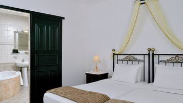 Habitación de hotel con cama, mesita de noche con lámpara, baño con bañera y lavabo, y una puerta oscura.