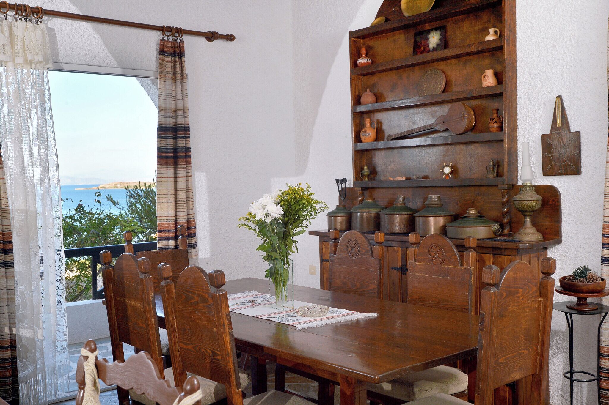 Un comedor con una mesa de madera, sillas, un florero con flores y una estantería con utensilios de cocina.