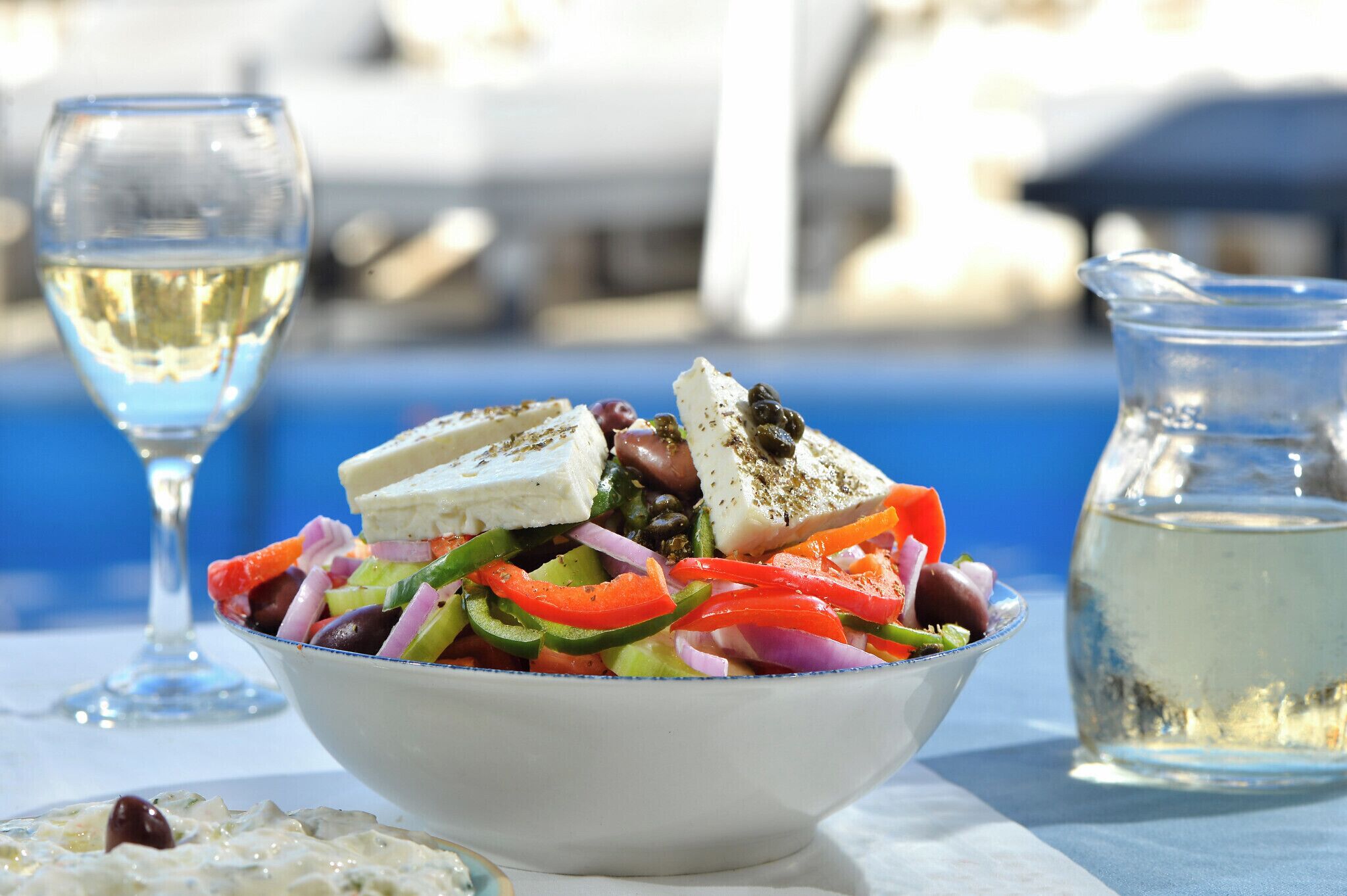 Un tazón de ensalada con queso feta, aceitunas y verduras, acompañado de una copa de vino blanco y una jarra de agua sobre una mesa.