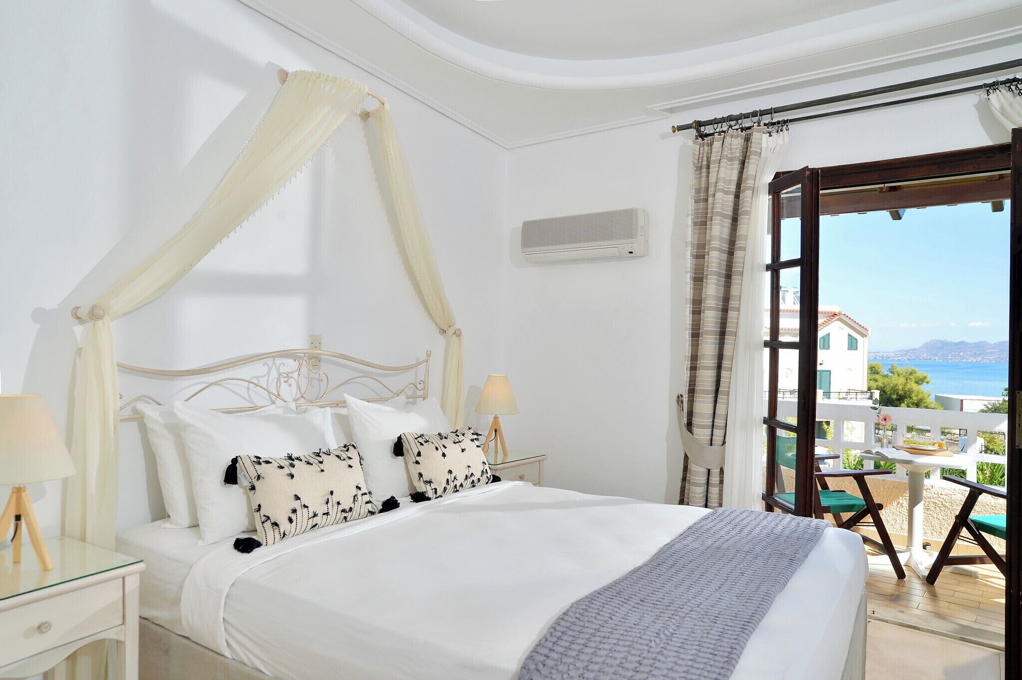 A bedroom with a four-poster bed, a bedside table with a lamp, and a view of the sea through an open door.