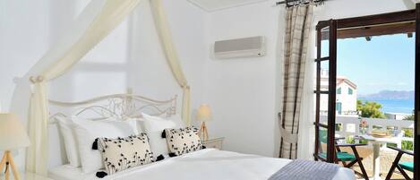 A bedroom with a four-poster bed, a bedside table with a lamp, and a view of the sea through an open door.