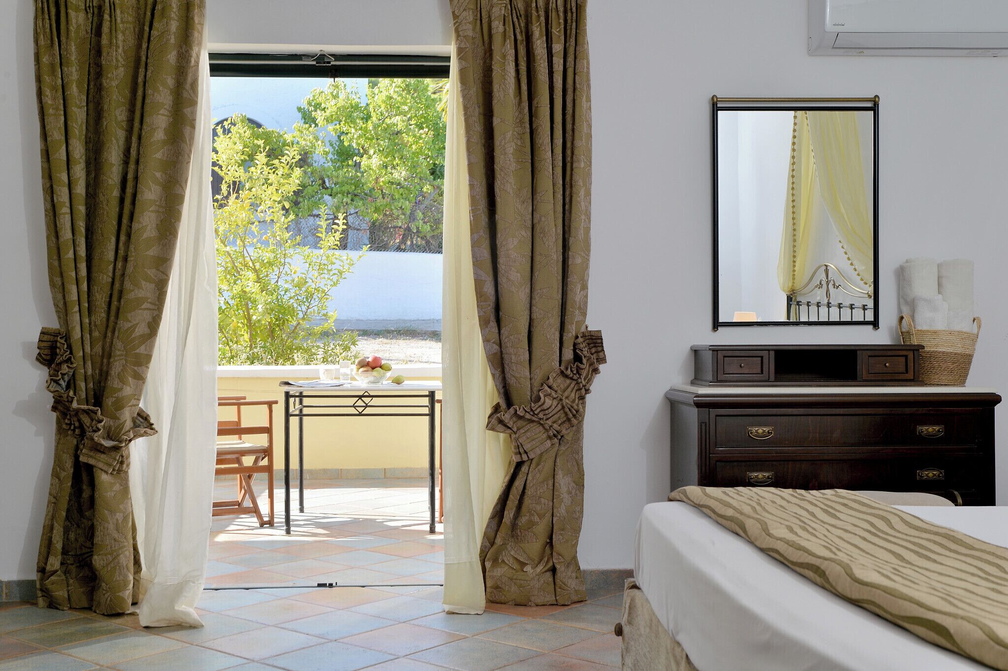 A room with a bed, a dresser, a mirror, and a view of a balcony with a table and chairs.