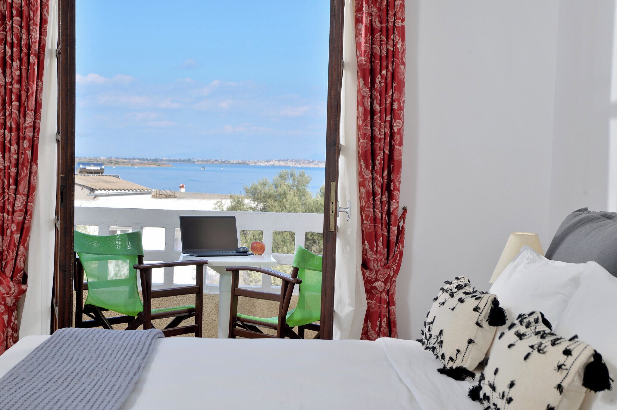 Un dormitorio con una cama, un ordenador portátil sobre una mesita pequeña, dos sillas y vistas al mar a través de cortinas abiertas.