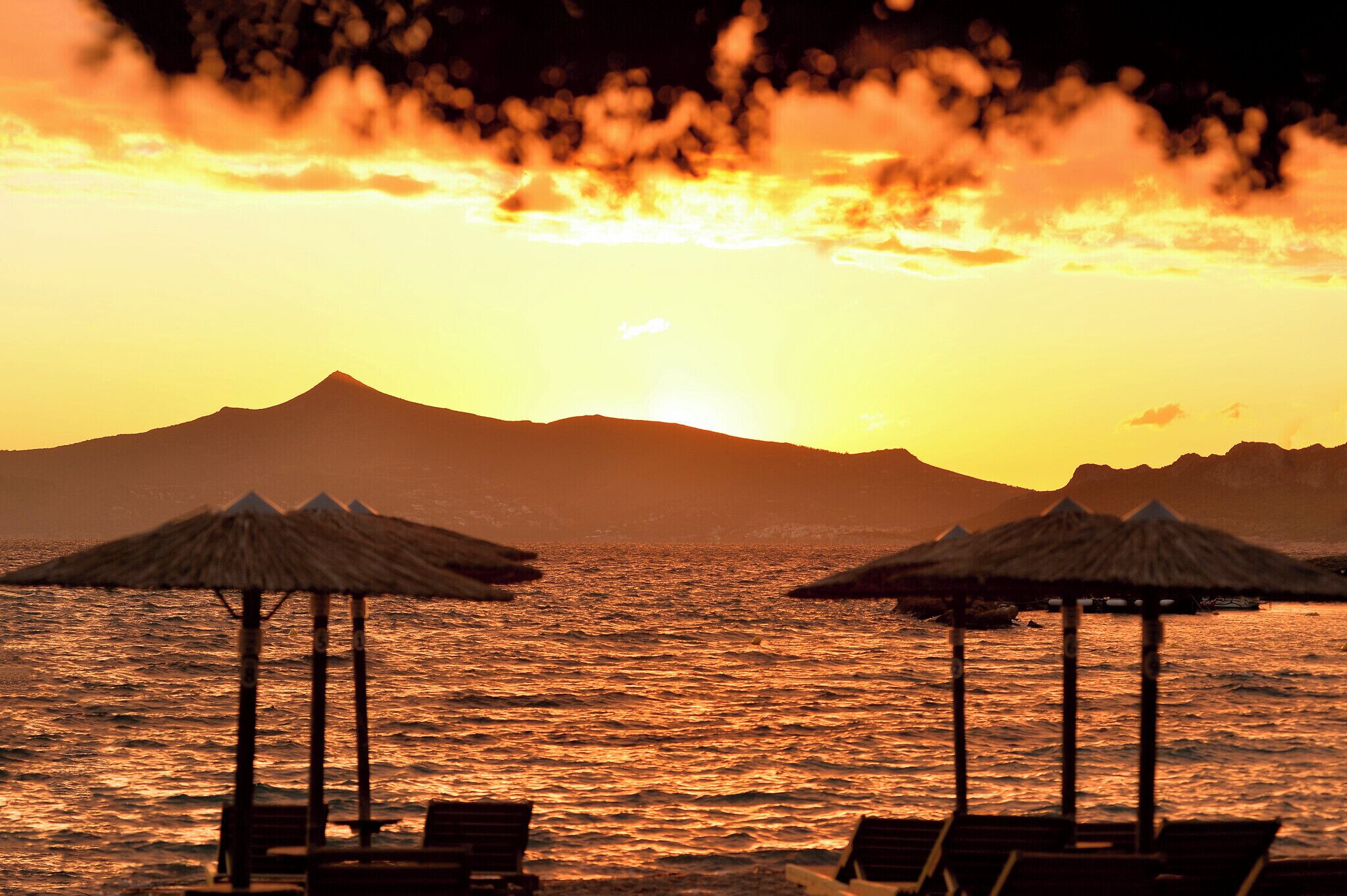 Escena de playa con sombrillas de paja y sillas de descanso, un atardecer y una montaña al fondo.