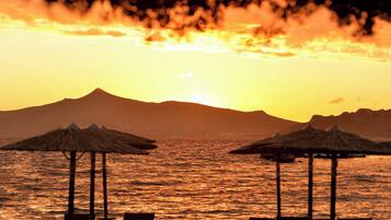 Escena de playa con sombrillas de paja y sillas de descanso, un atardecer y una montaña al fondo.