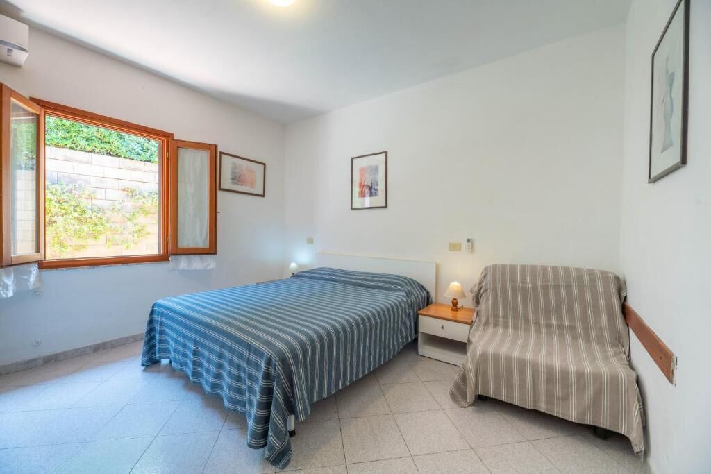 A room with two beds, a chair, and a window with a view of greenery.
