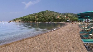 A pebbled beach with a clear sky and a distant hill with buildings. - Casa Luppoli (Portoferraio)