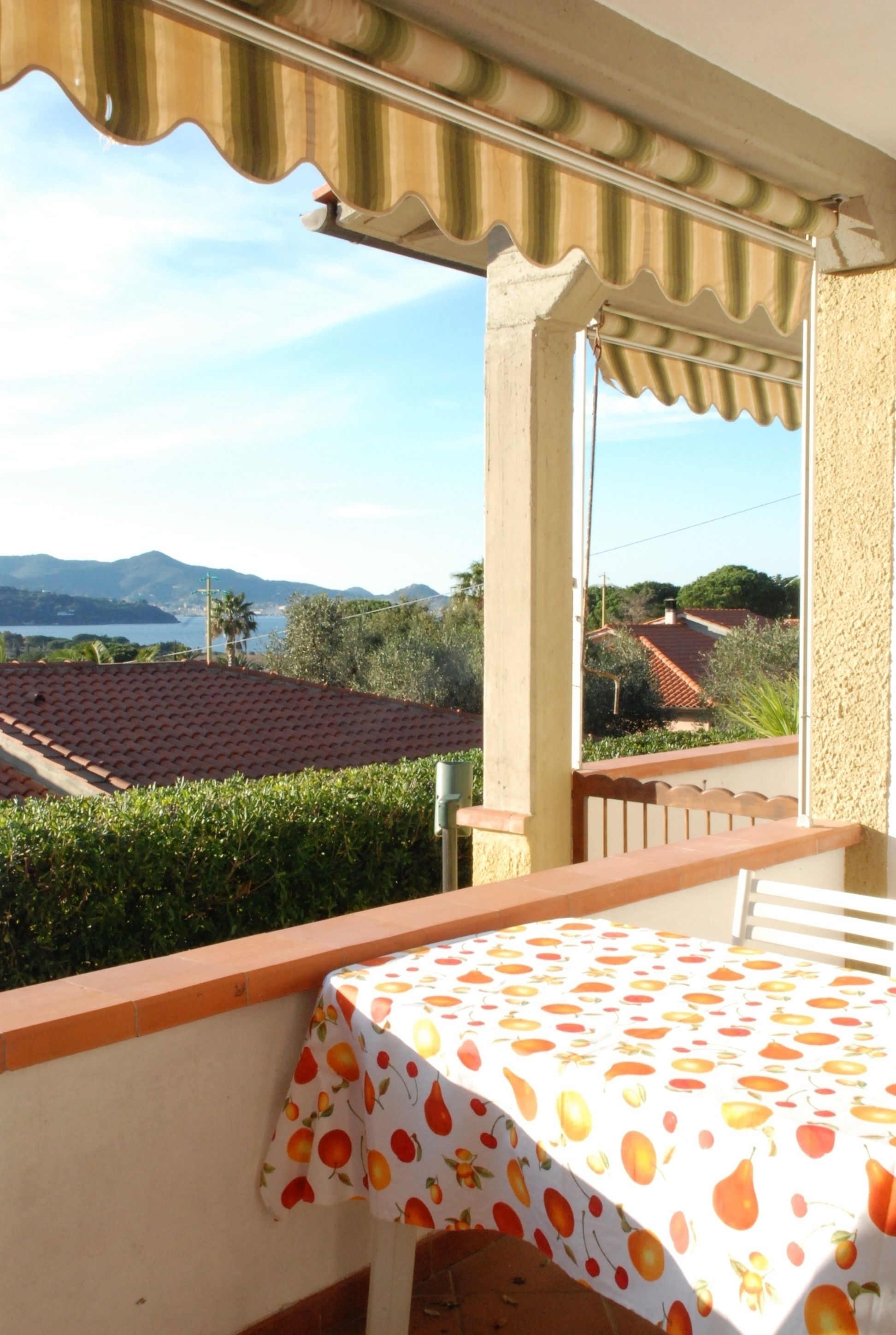 A balcony with a table covered in a fruit-patterned tablecloth, overlooking a coastal landscape with houses and mountains.