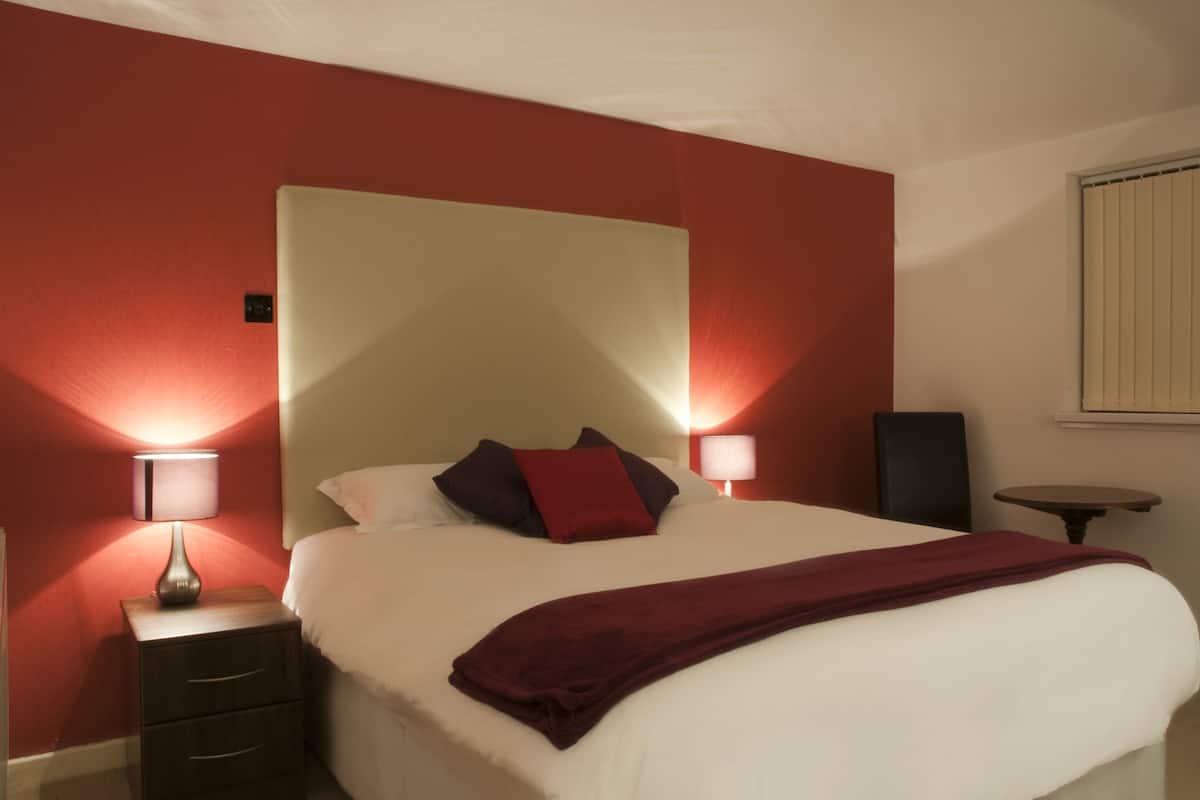 A hotel room with a large bed, two bedside tables with lamps, a small table, and a window with blinds.