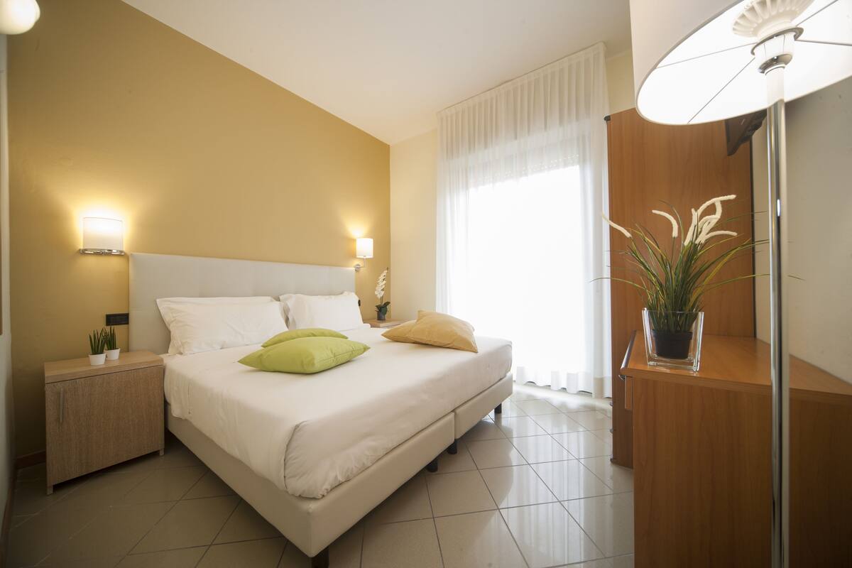 A neatly arranged bedroom with a bed, bedside tables, a nightstand, a potted plant, and a mirror.
