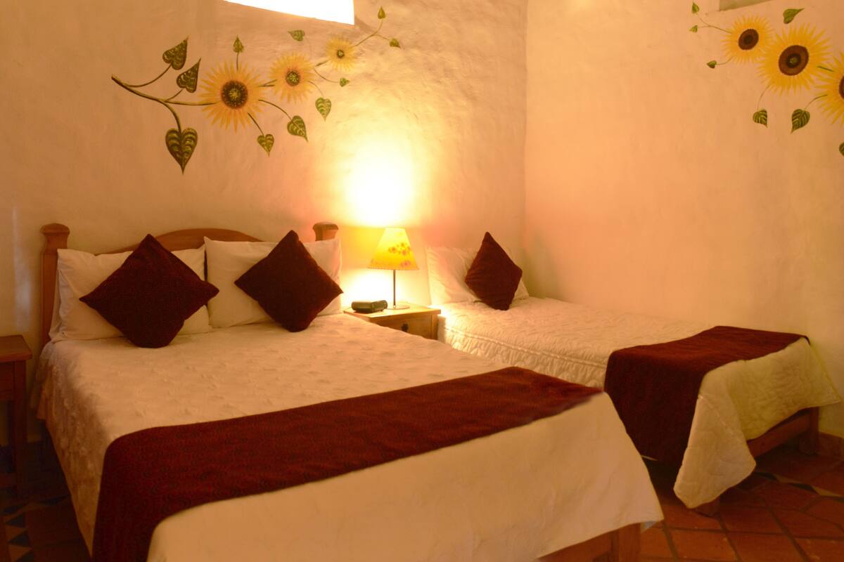 A room with two beds, each with a red throw blanket and white bedding. There is a nightstand with a lamp between the beds. The walls are decorated with sunflower and leaf patterns.