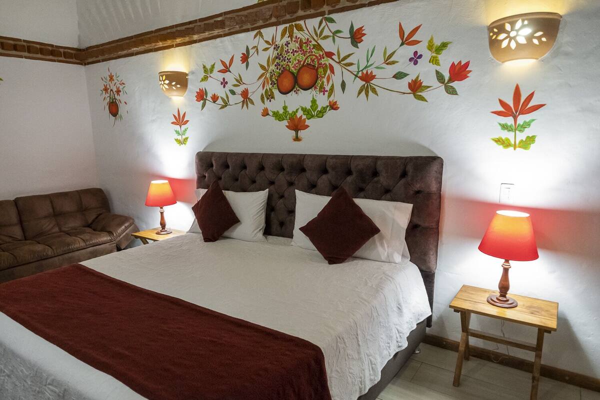 A bedroom with a large bed, two bedside tables with lamps, a brown leather sofa, and a decorative wall mural.