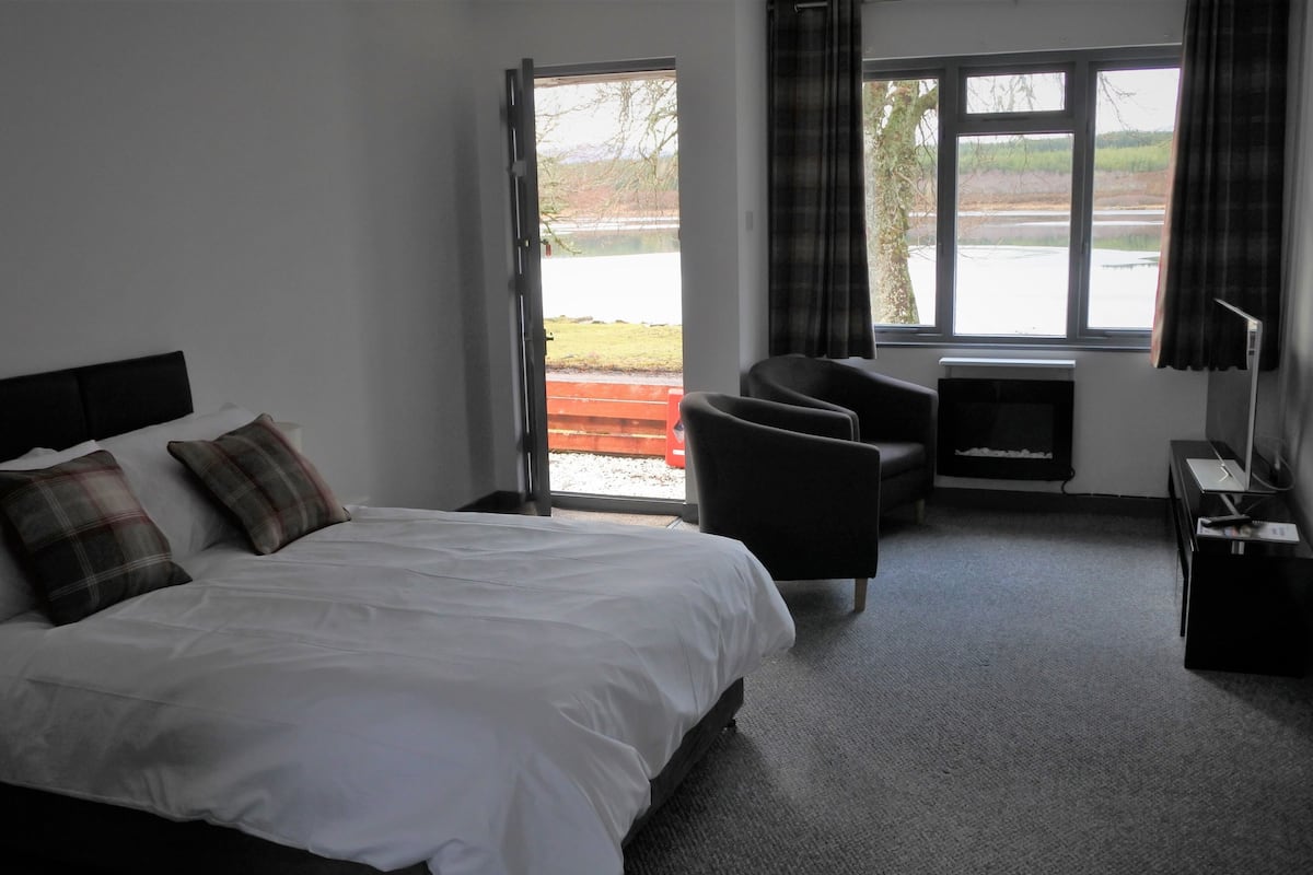 A modern hotel room with a large bed, a desk, a chair, and a view of a lake through the window.