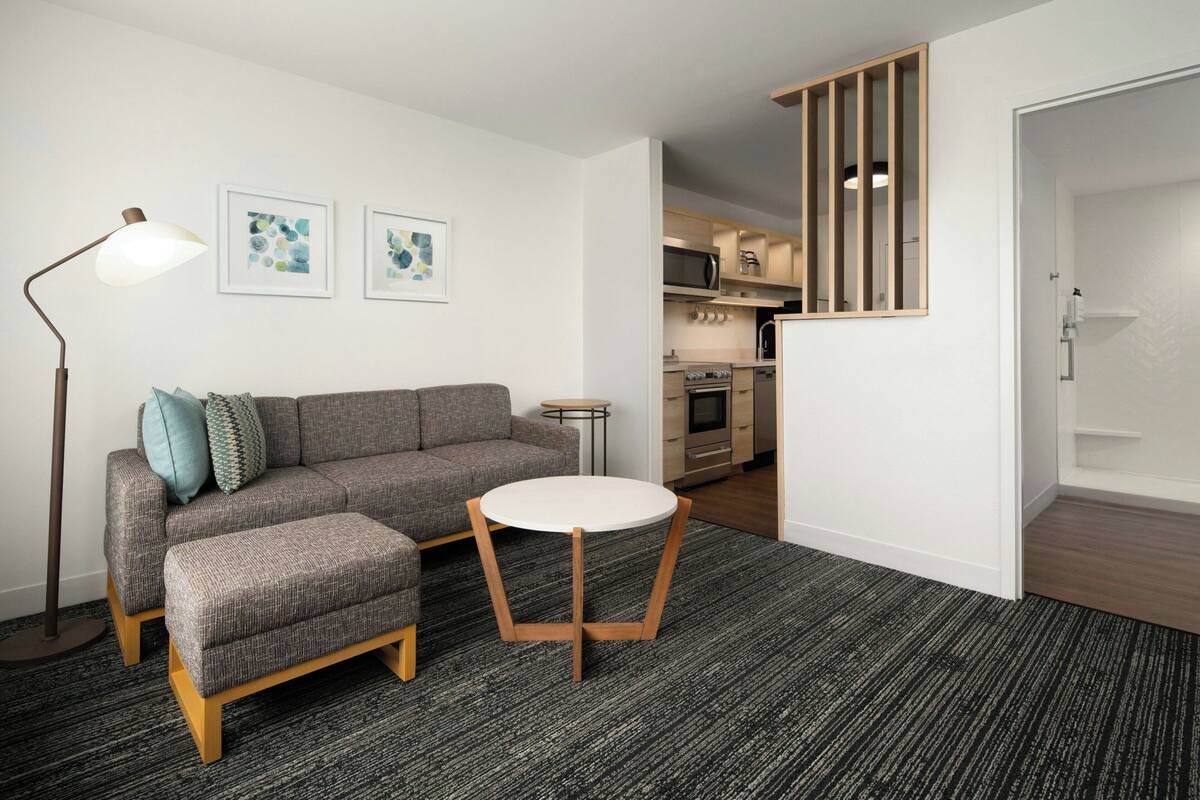 A modern living room with a grey sofa, a round coffee table, and a floor lamp. There is a kitchen visible in the background with wooden cabinets and a microwave.