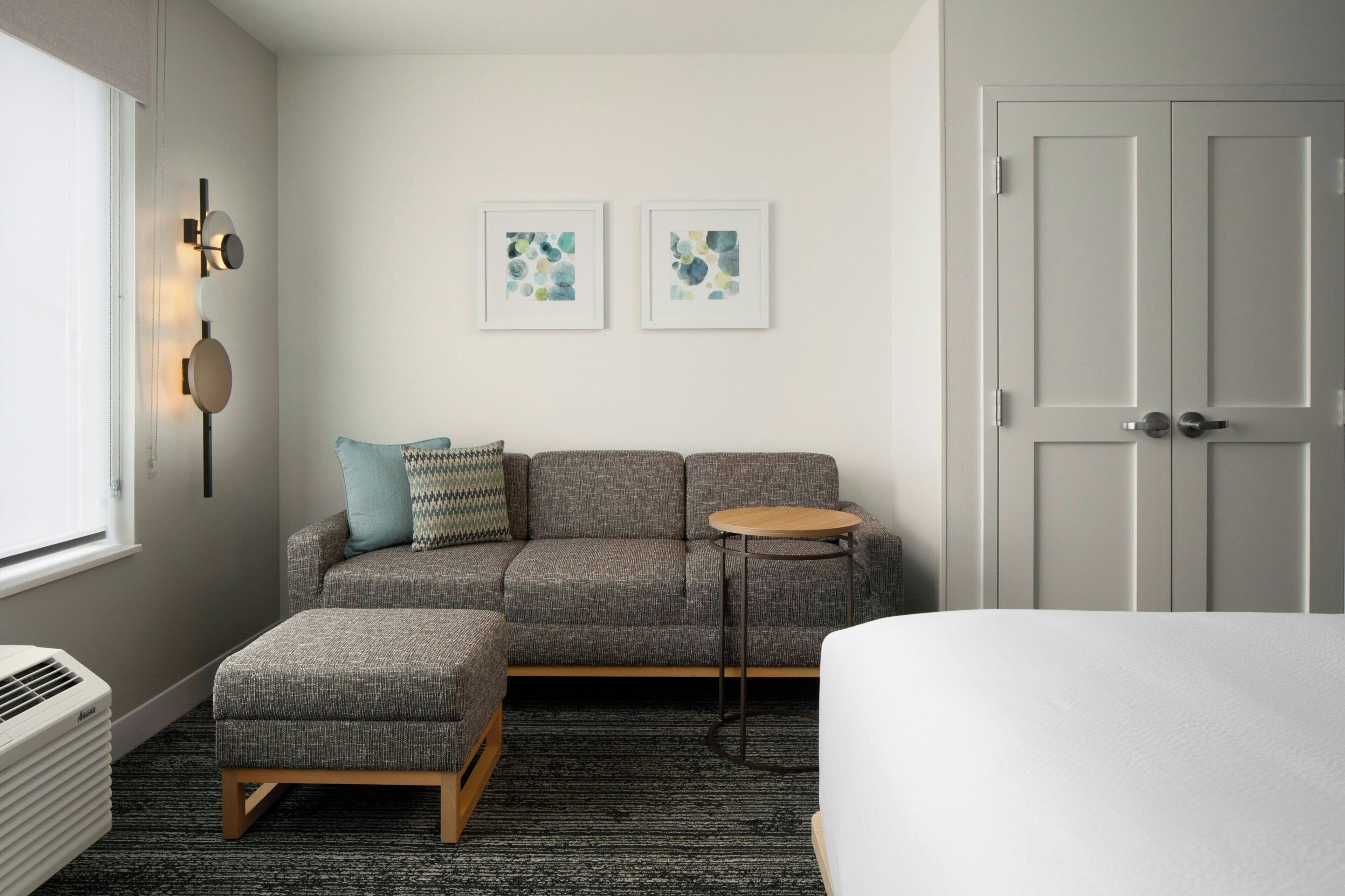 Una habitación de hotel moderna con un sofá gris, una mesa redonda de madera, una cama blanca y un radiador. Se observan dos obras de arte enmarcadas en la pared y una puerta a la derecha.