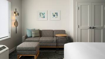 Una habitación de hotel moderna con un sofá gris, una mesa redonda de madera, una cama blanca y un radiador. Se observan dos obras de arte enmarcadas en la pared y una puerta a la derecha.