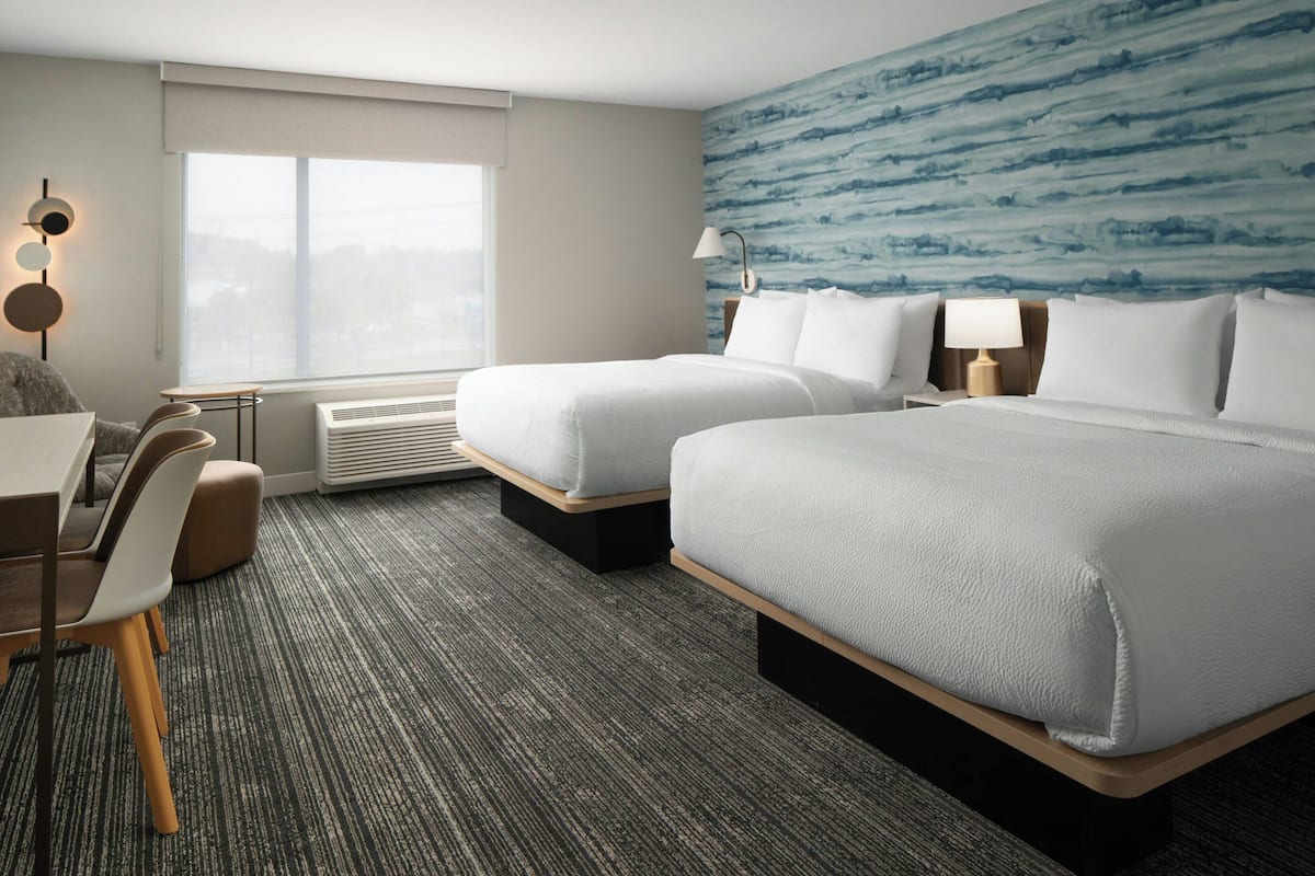 A hotel room with two beds, a desk, chairs, a window, and a wall with a blue pattern.