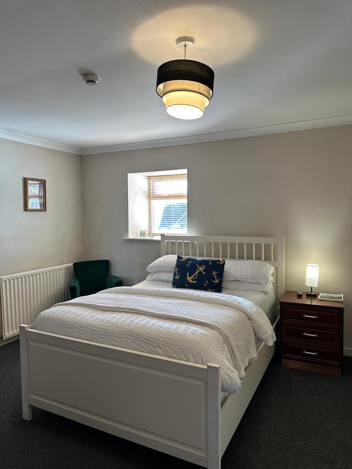 A bedroom with a white bed, a nightstand, a window with blinds, a ceiling light, and a radiator.